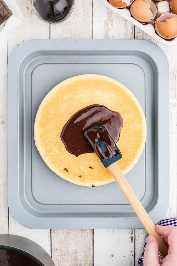 A hand spreads chocolate frosting on a round cake with a spatula, eggshells are nearby.