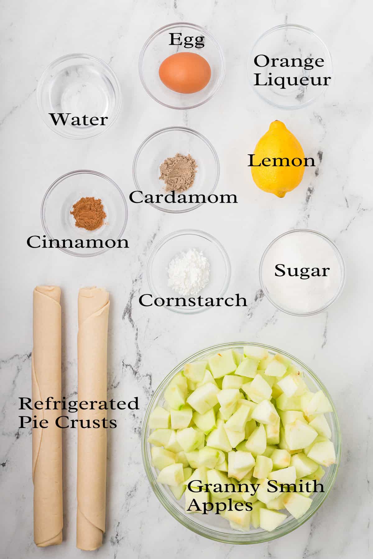 Ingredients for apple cobbler arranged on a white marble surface, including two pie crusts, a bowl of Granny Smith apple chunks, an egg, lemon, orange liqueur, water, cinnamon, cardamom, cornstarch, and sugar.