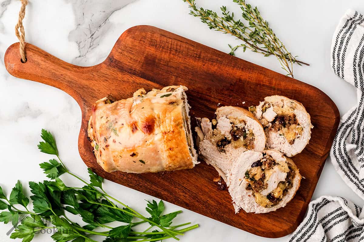 Sliced stuffed turkey roulade on a wooden cutting board with herbs and a striped kitchen towel nearby.