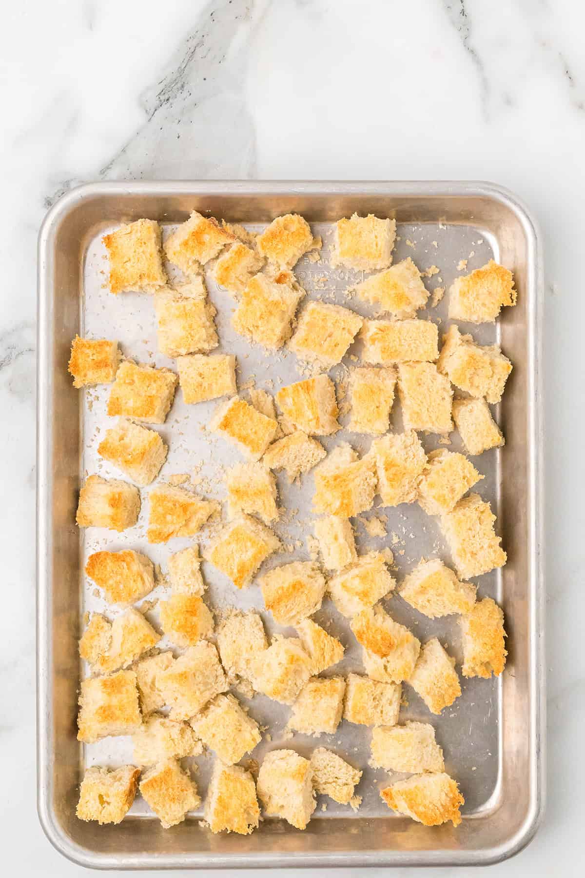 Cubed pieces of bread are scattered on a metal baking sheet set on a white marble surface.