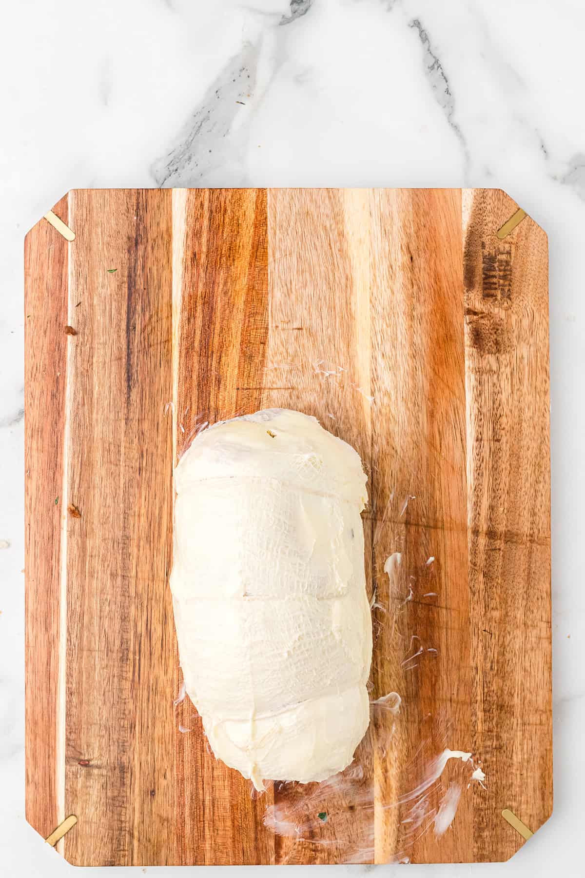 A butter covered, rolled and stuffed turkey breast sits on a wooden cutting board on a white marble surface.