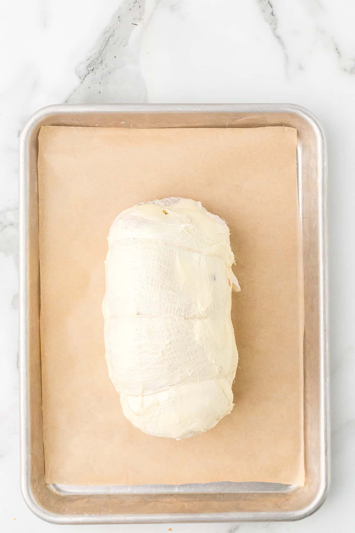 A rolled, stuffed turkey breast covered in butter sits on parchment paper on a metal baking sheet, on a marble surface.