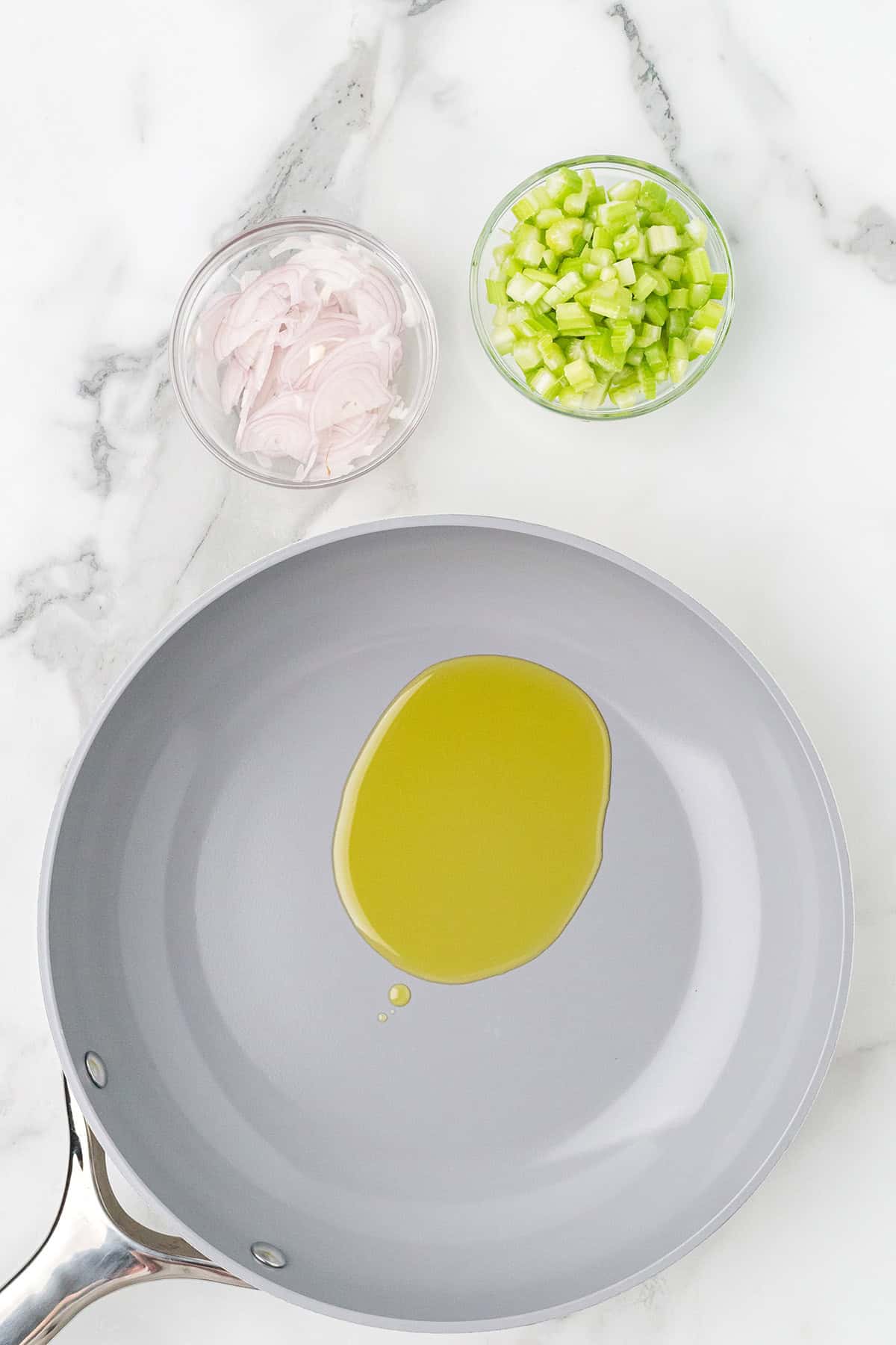 A frying pan with olive oil, next to bowls of sliced shallots and diced celery on a marble surface.