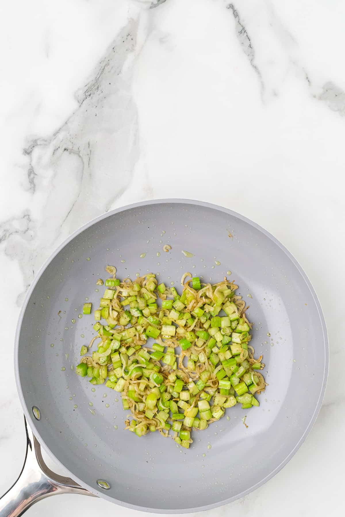 Chopped celery and shallots sautéing in a gray nonstick skillet on a marble countertop.