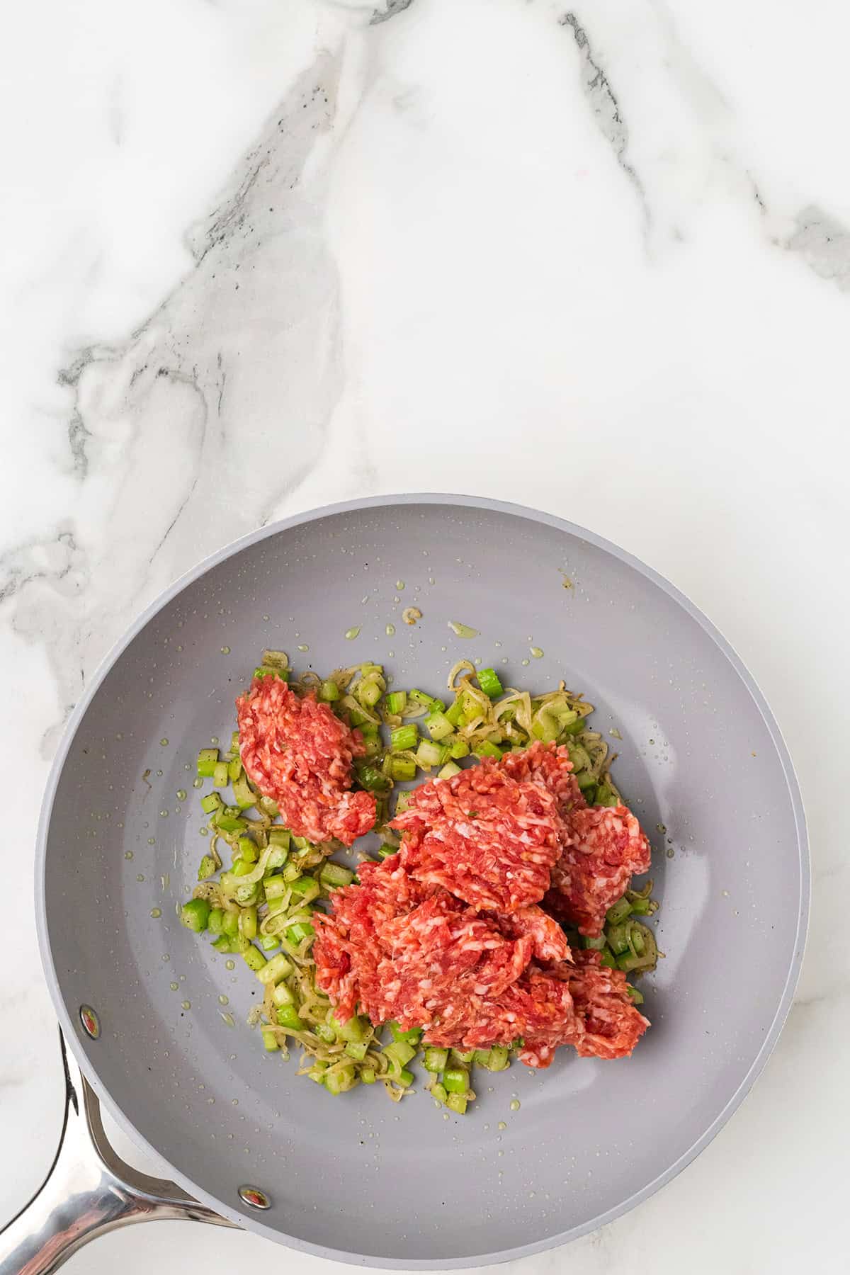 Italian sausage and chopped green vegetables cooking in a gray skillet on a marble countertop.