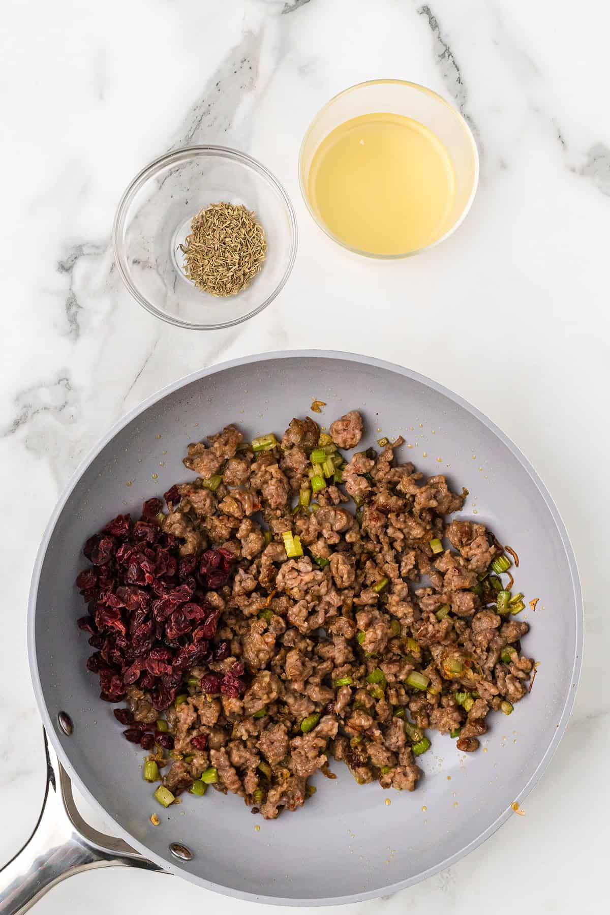 Italian sausage, dried cranberries, and diced celery in a skillet. Small bowls of broth and herbs beside it.