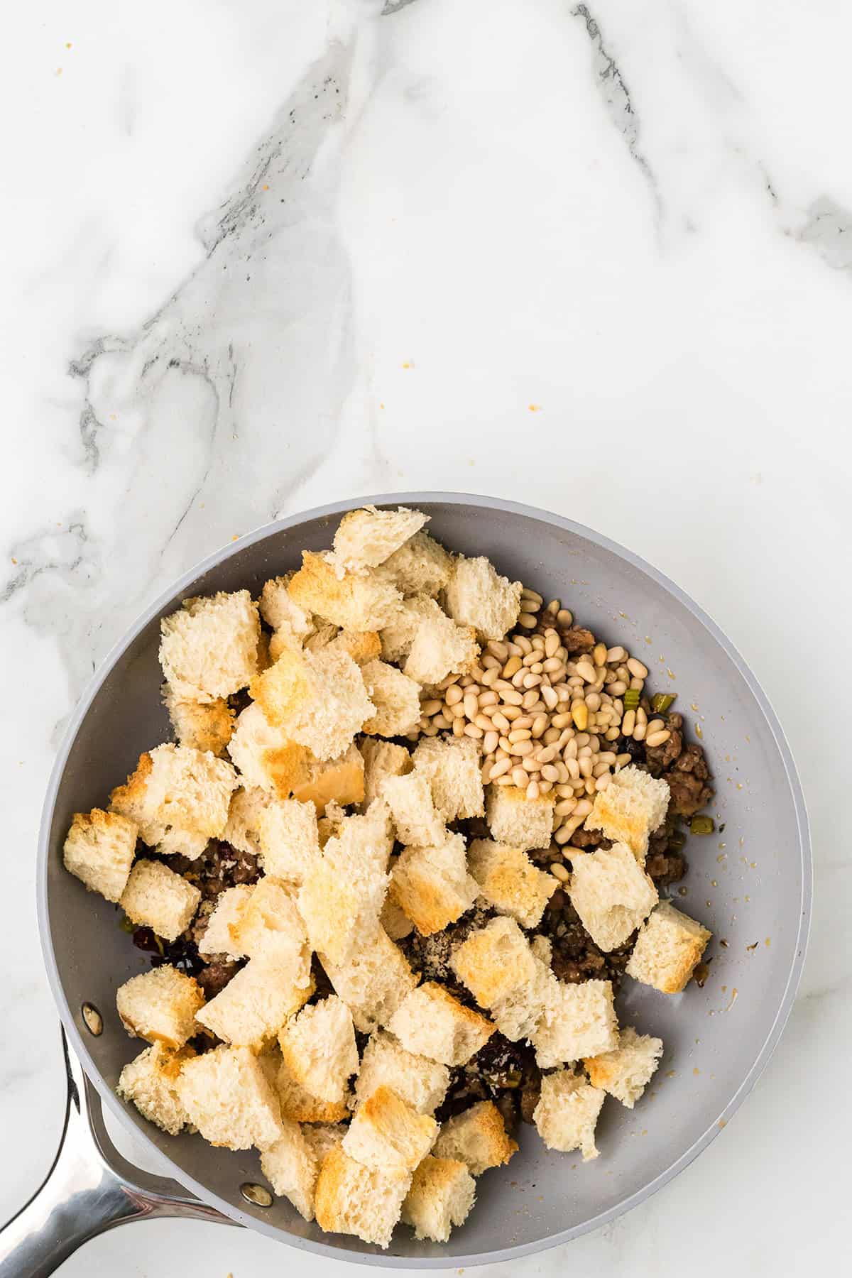 A frying pan with bread cubes, pine nuts, and a dark mixture on a marble countertop.