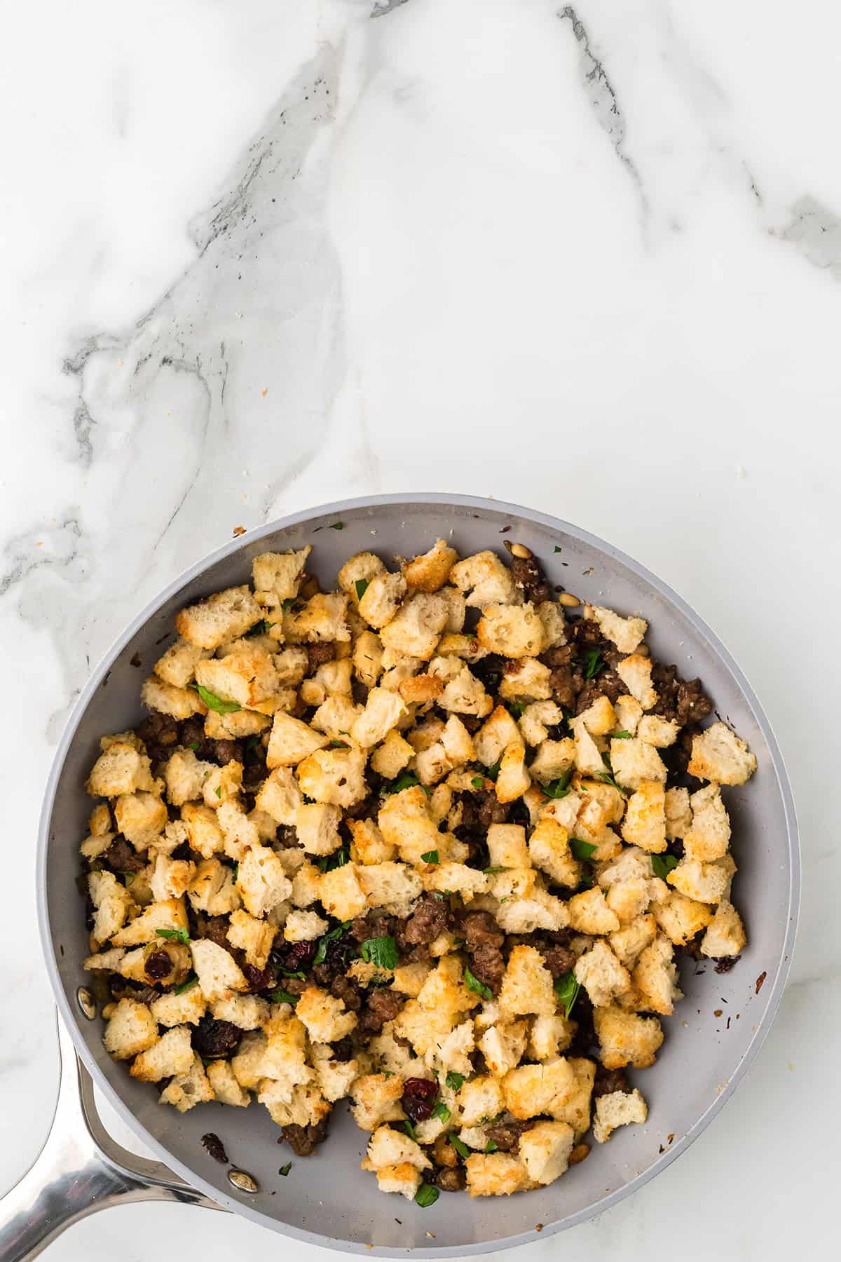A pan of cooked stuffing with bread cubes, herbs, and sausage on a marble countertop.