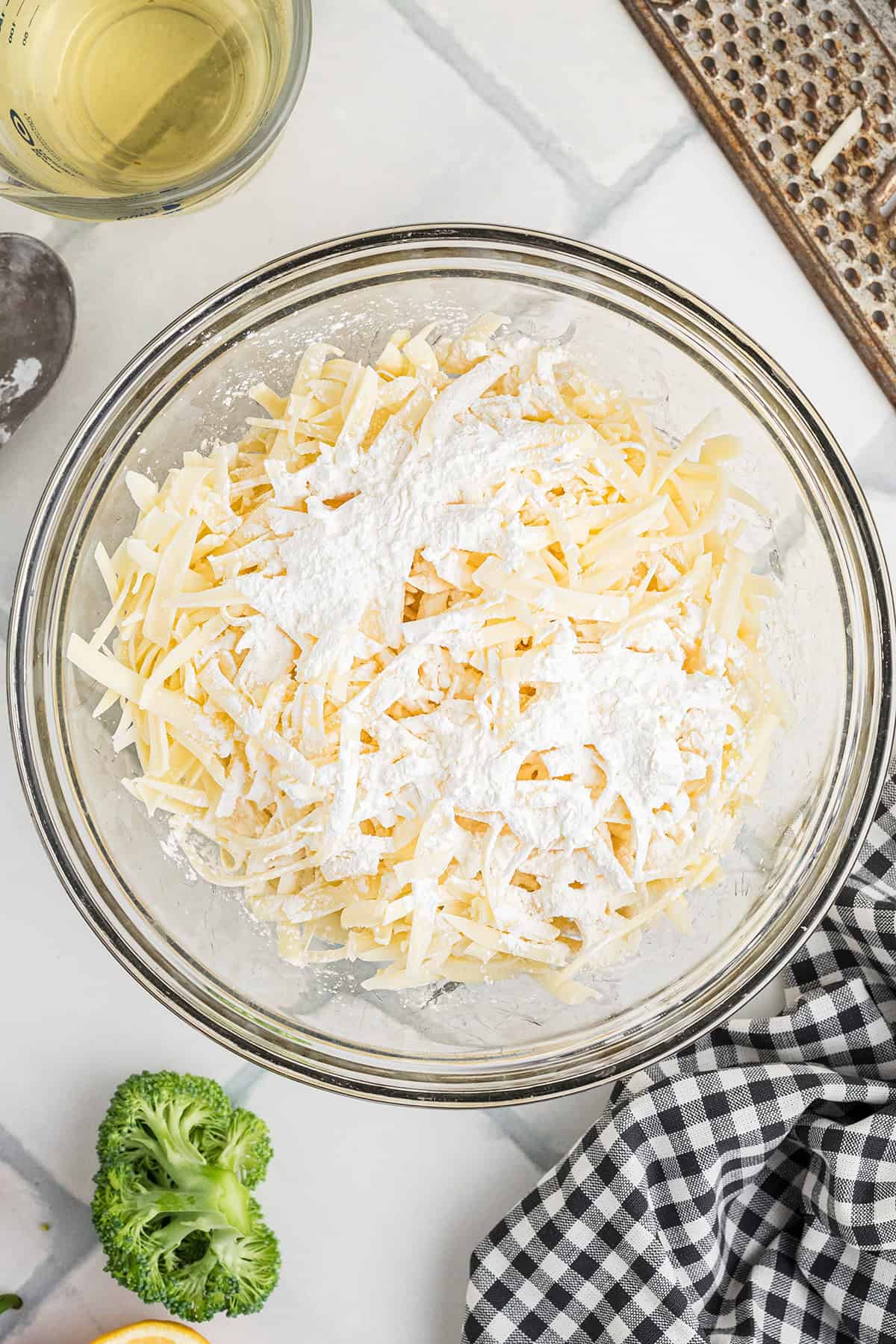 A glass bowl with shredded cheese and cornstarch on a countertop, surrounded by ingredients and a checkered cloth.