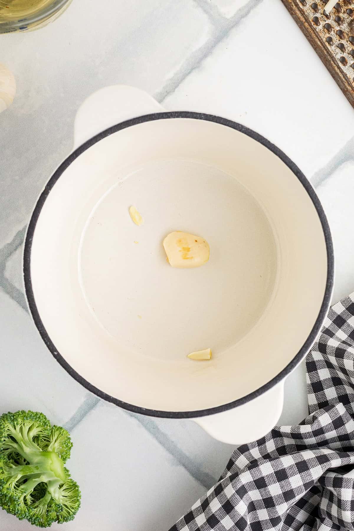White pot with a slice of garlic inside, next to broccoli and a black-and-white checkered cloth on a countertop.