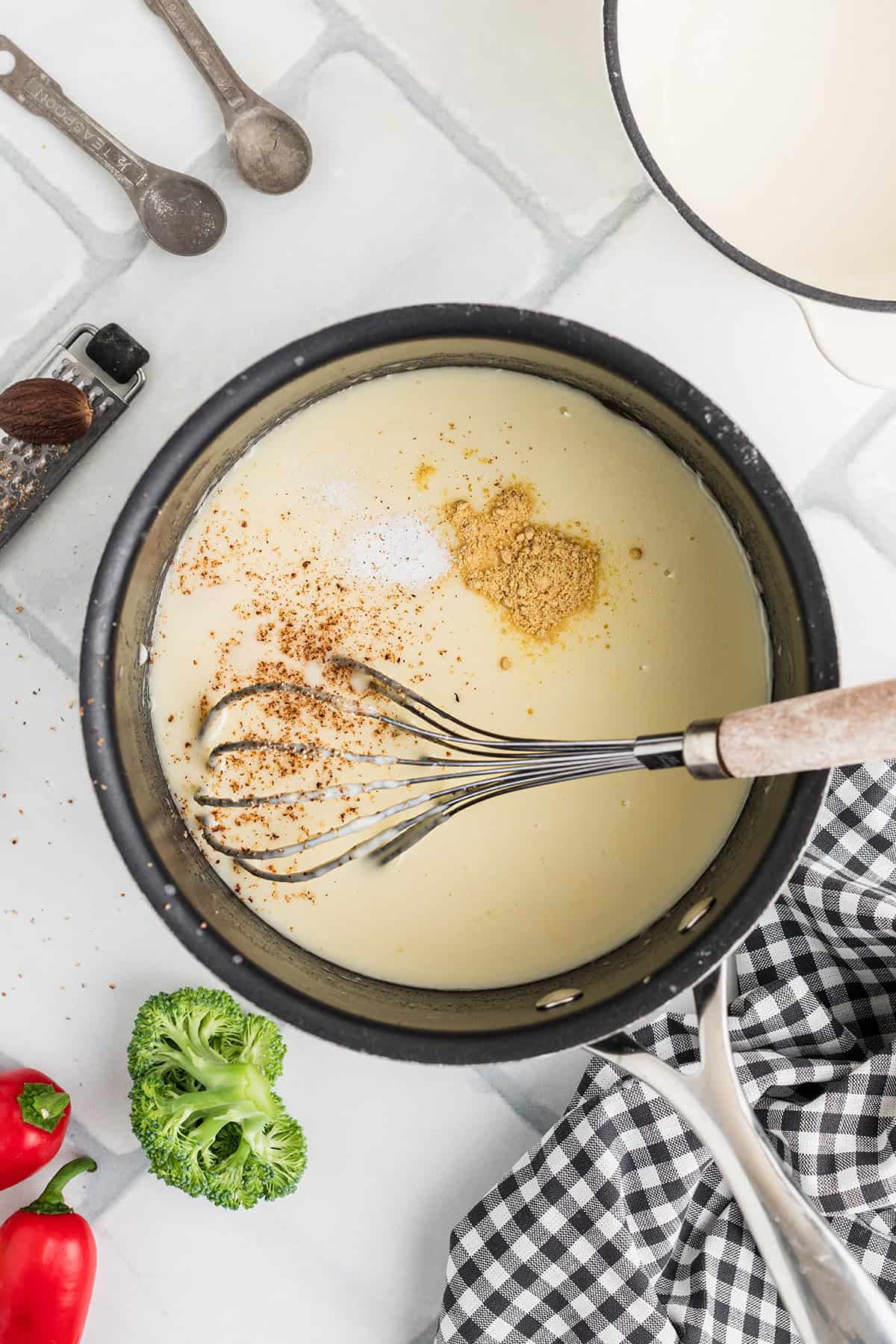 A saucepan with creamy sauce, spices, and a whisk, next to broccoli, pepper, and a checked cloth on a counter.