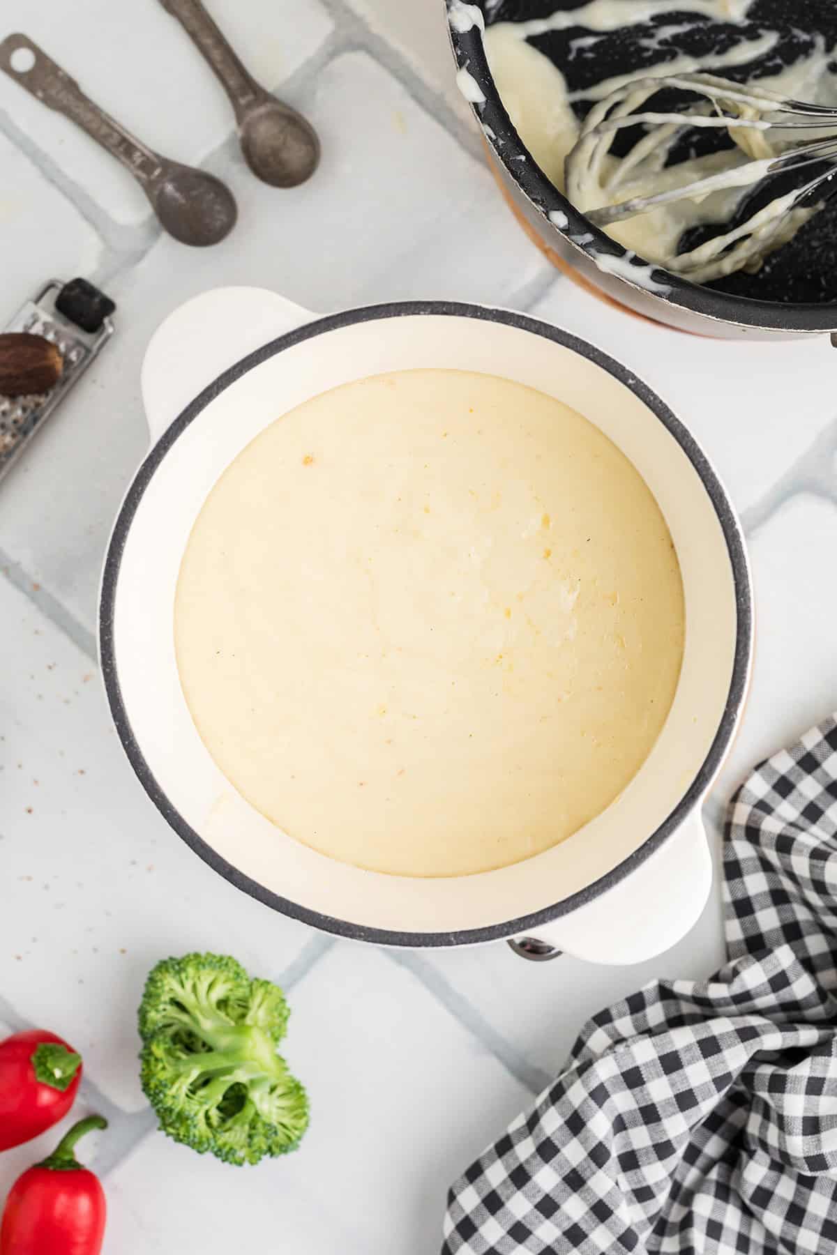 Overhead view of a pot filled with creamy fondue, surrounded by broccoli, red pepper, and kitchen utensils.
