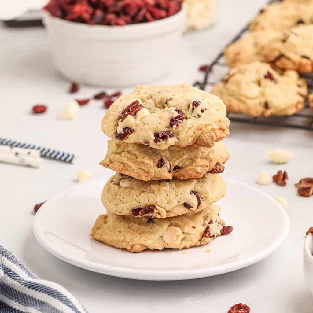 Four cranberry and white chocolate cookies stacked on a white plate, with more cookies and ingredients in background.