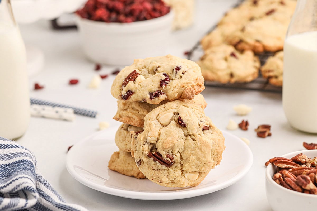 Three white chocolate cranberry cookies stacked on a plate, with milk bottles and more cookies in the background.