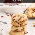 A stack of cranberry pecan cookies on a white plate with more cookies and nuts in the background.