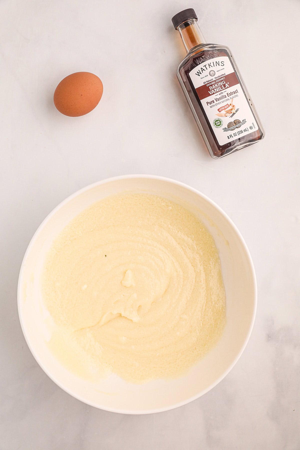 A bowl of creamed mixture, a whole egg, and a bottle of vanilla extract on a white surface.