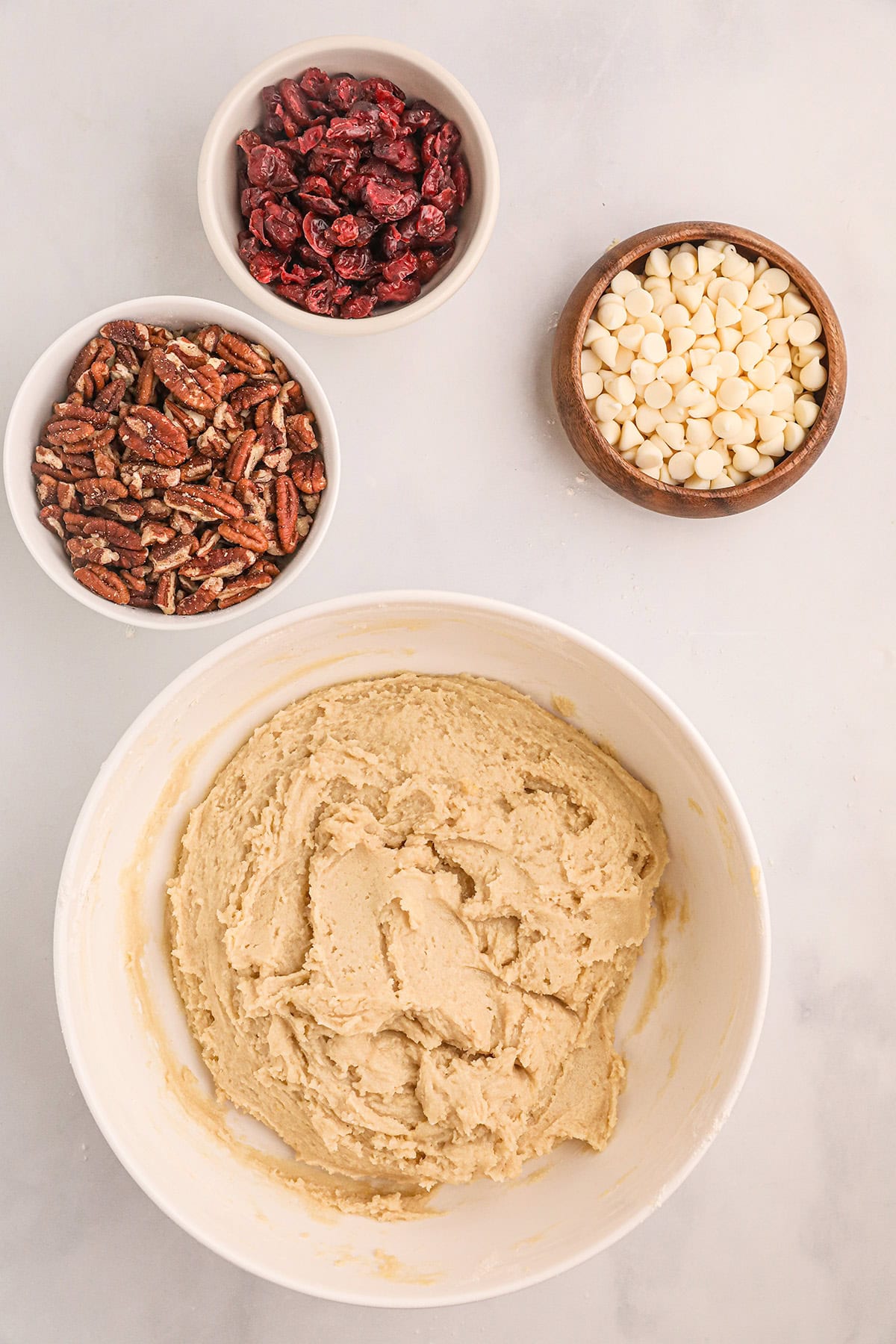 A bowl of cookie dough beside small bowls of dried cranberries, pecans, and white chocolate chips.