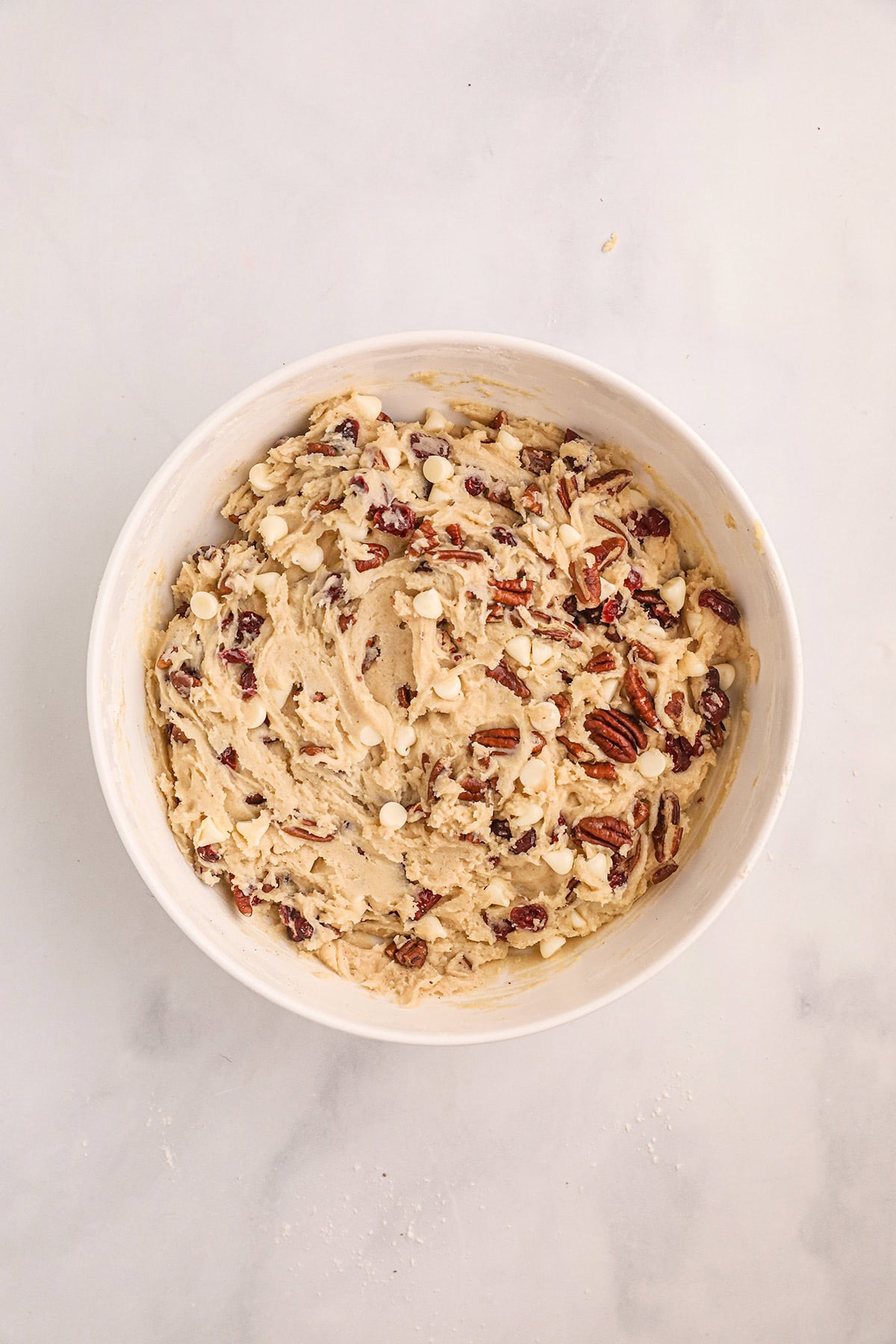 A bowl of cookie dough with white chocolate chips, pecans, and dried cranberries on a light surface.