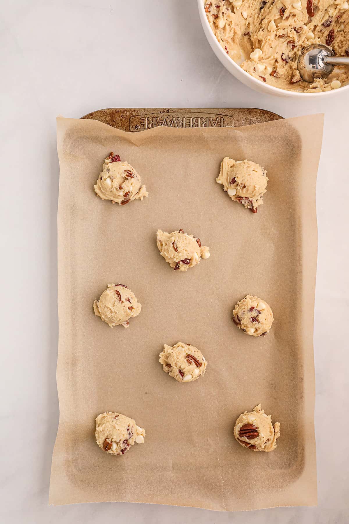 Eight scoops of cookie dough with nuts on parchment paper, ready to bake, beside a bowl of more dough.