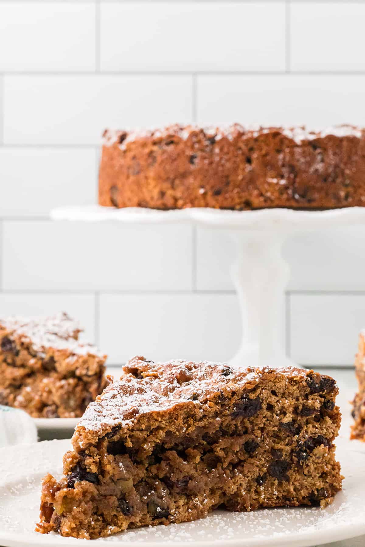 A slice of cake in the foreground with a cake on a pedestal in the background.