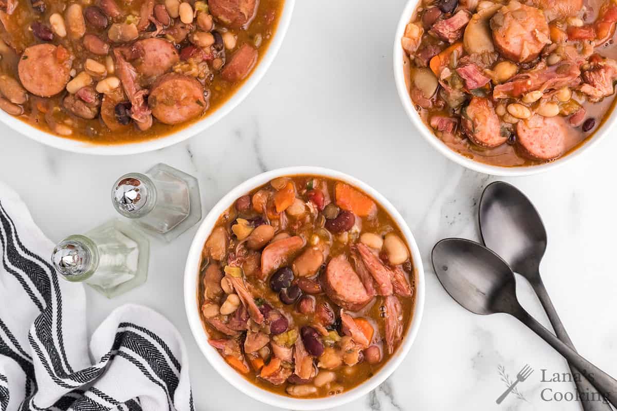 Three bowls of mixed bean soup on a white table with spoons, salt, pepper, and a striped cloth nearby.