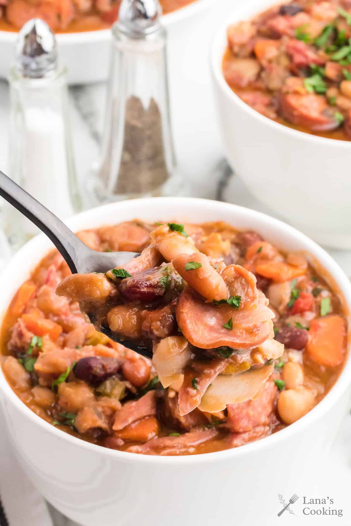 A spoonful of hearty bean and sausage stew held above a bowl, with salt and pepper shakers in the background.
