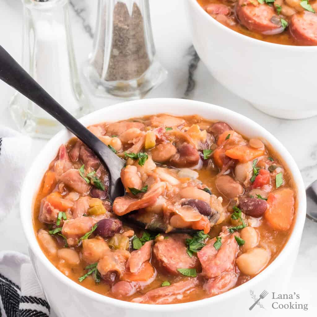 A bowl of bean and sausage stew with a spoon, garnished with herbs, next to salt and pepper shakers.