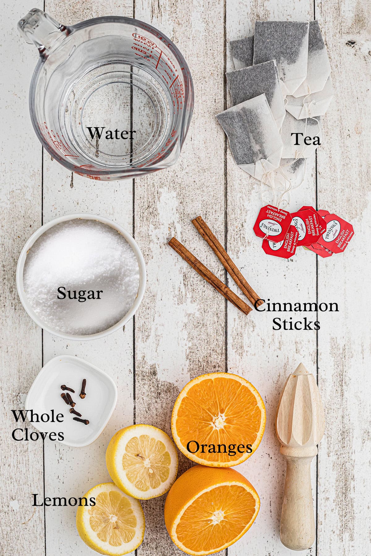 Top-down view of water, tea bags, sugar, cinnamon sticks, whole cloves, oranges, and lemons on a white surface.