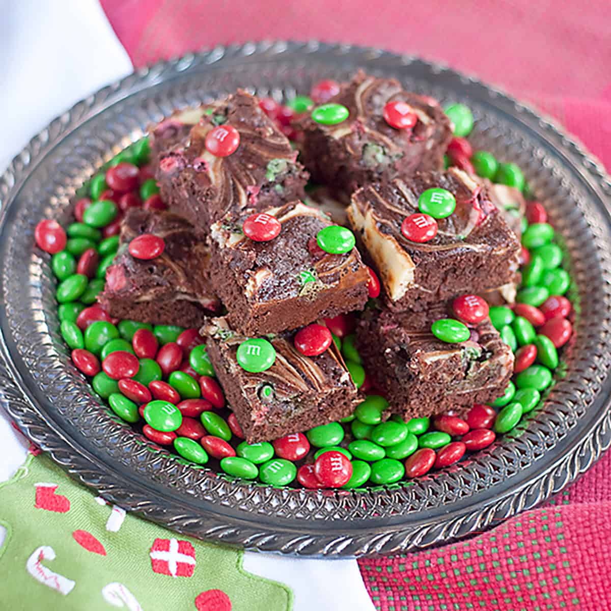 A plate of brownies topped with red and green M&M candies, surrounded by more M&Ms on a festive cloth.