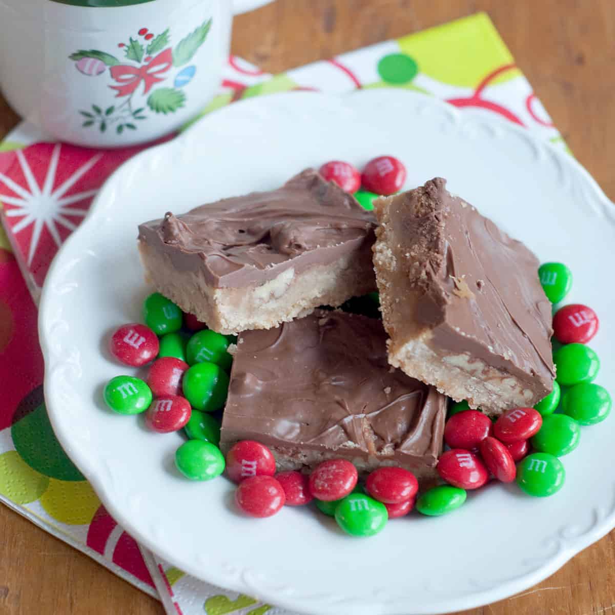 Three chocolate-covered bars on a plate surrounded by red and green M&M candies, with a holiday mug nearby.