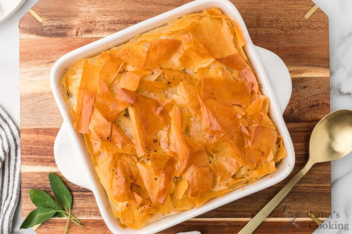 A golden, flaky phyllo pie in a white baking dish on a wooden board with a gold spoon nearby.