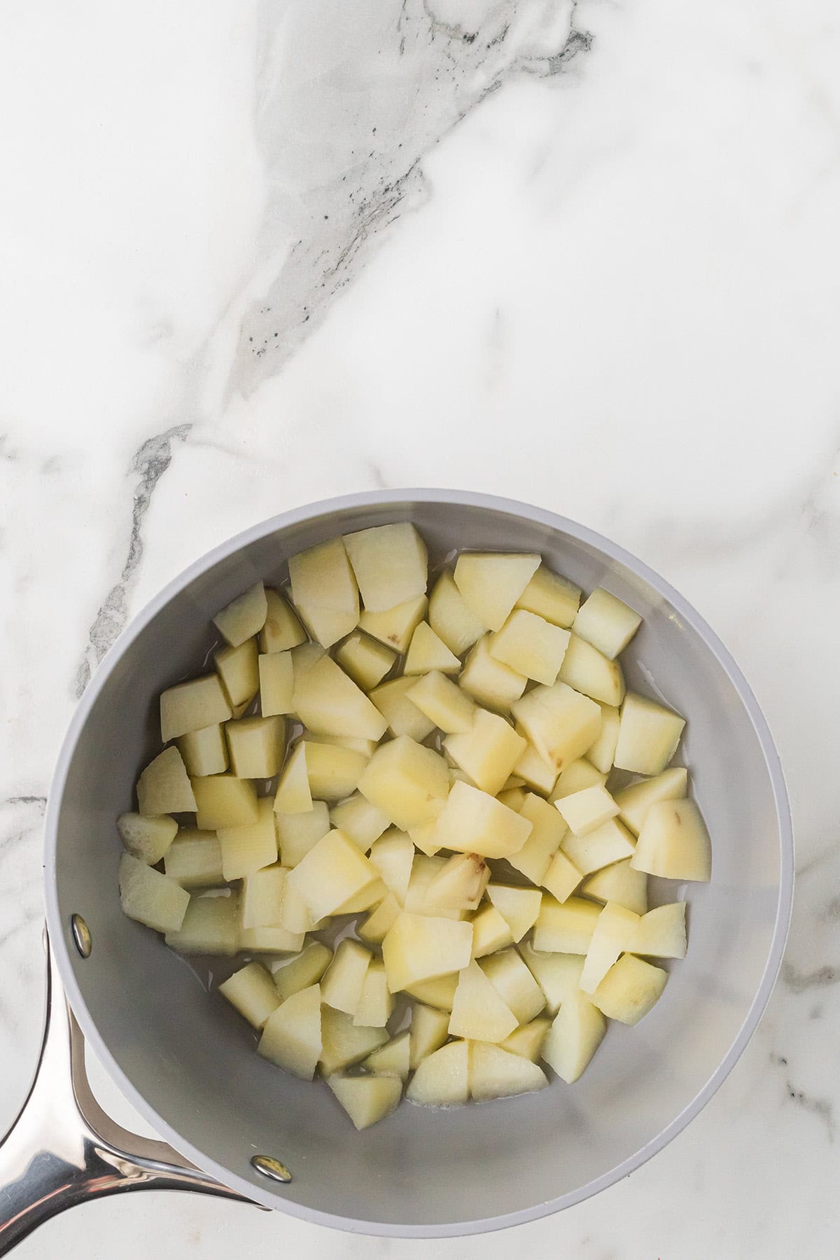 A saucepan filled with diced potatoes sits on a white marble countertop.