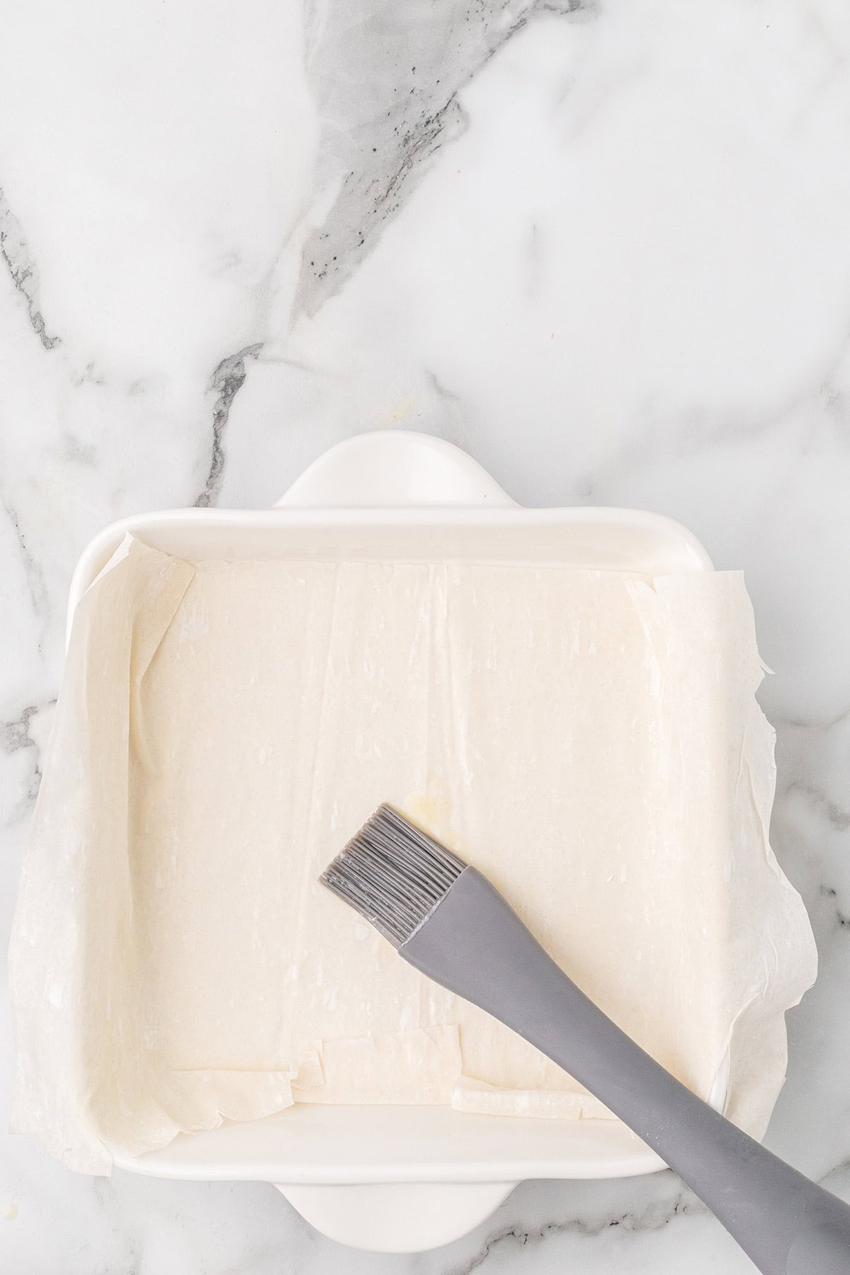 A pastry brush on layered phyllo dough in a square white baking dish, on a marble countertop.
