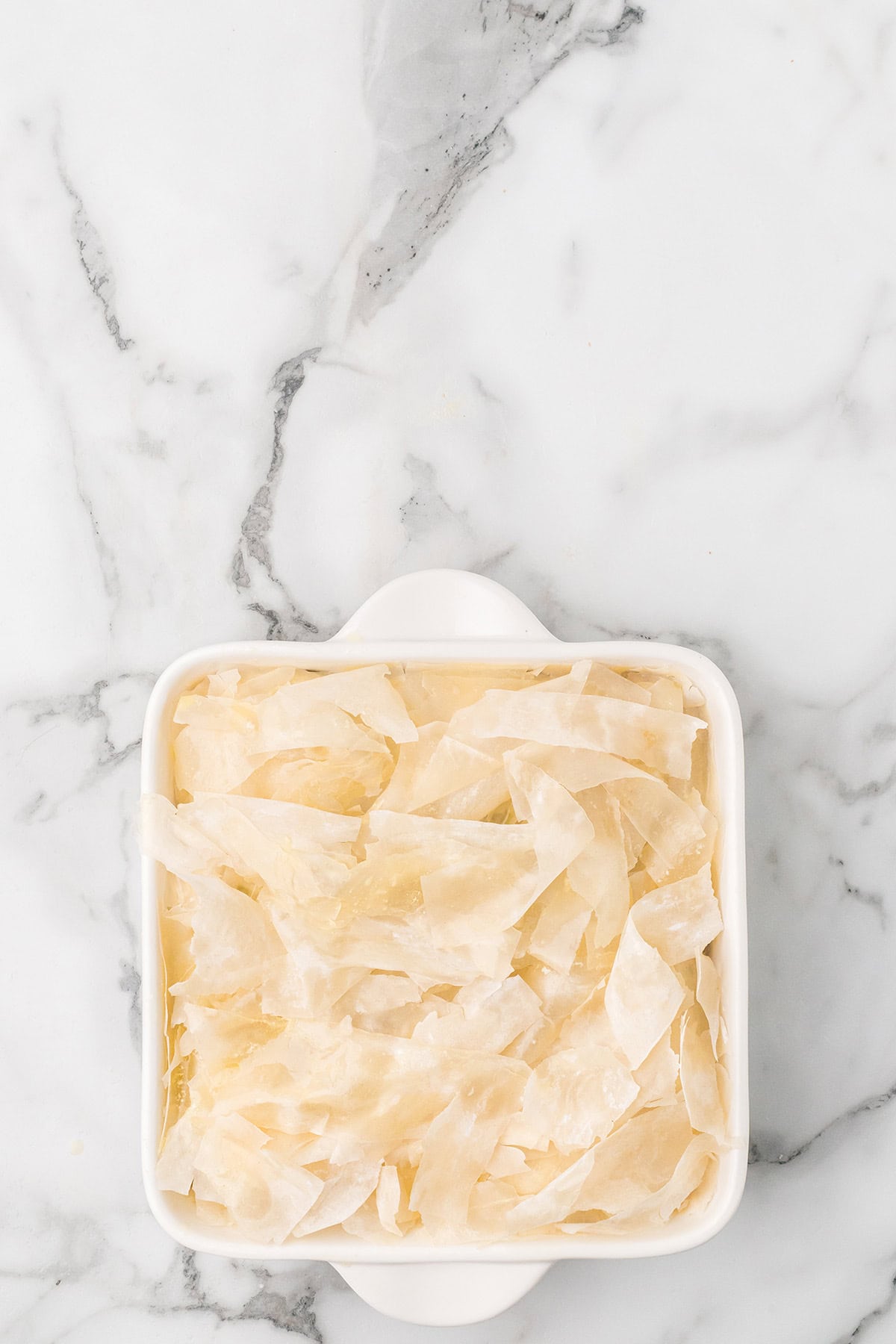 A white baking dish filled with uncooked phyllo dough on a marble surface.