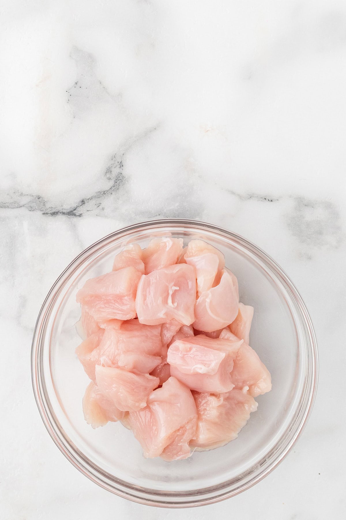 Glass bowl filled with raw, cubed chicken pieces on a white marble surface.