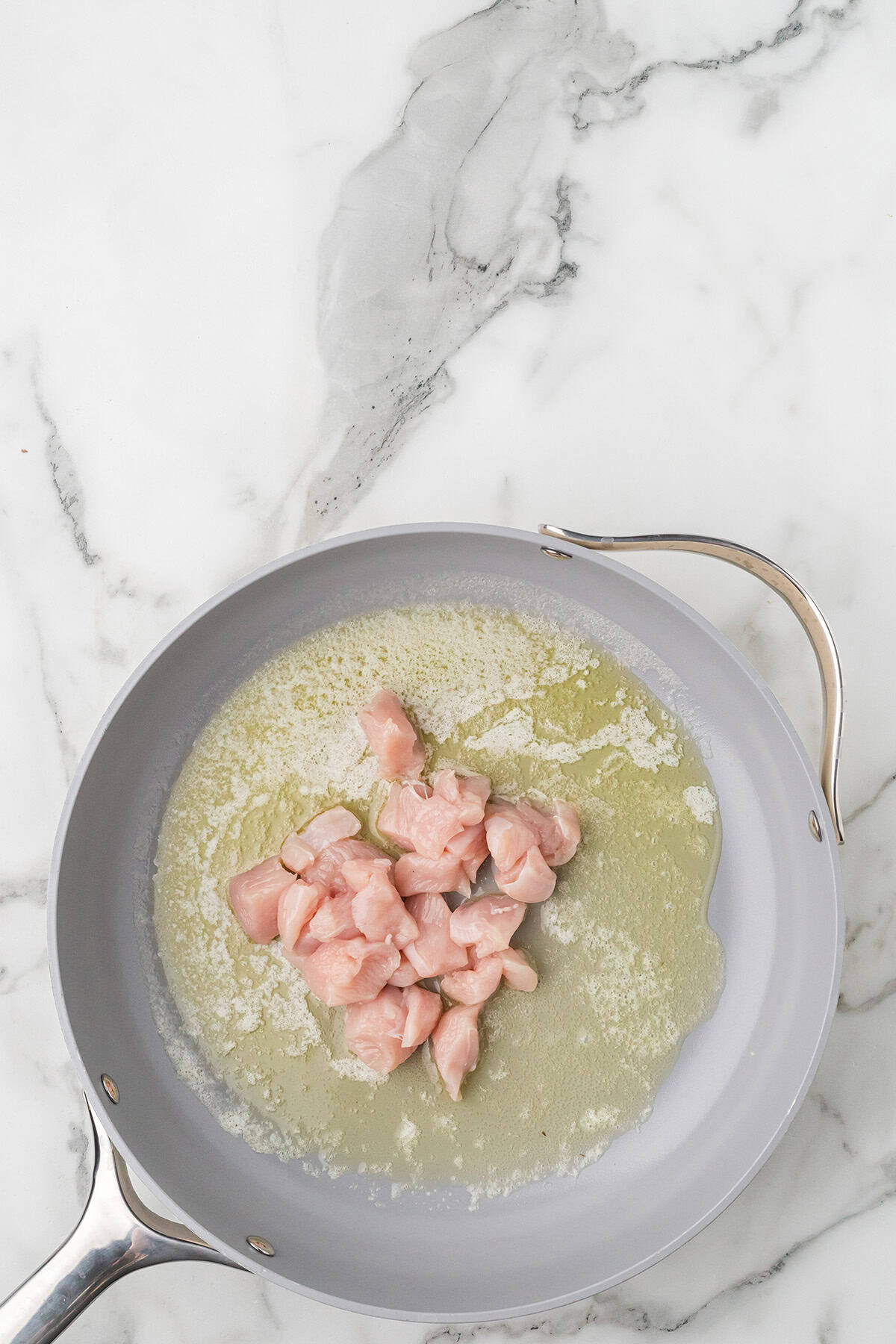 Raw chicken pieces cooking in a frying pan with melted butter on a white marble surface.