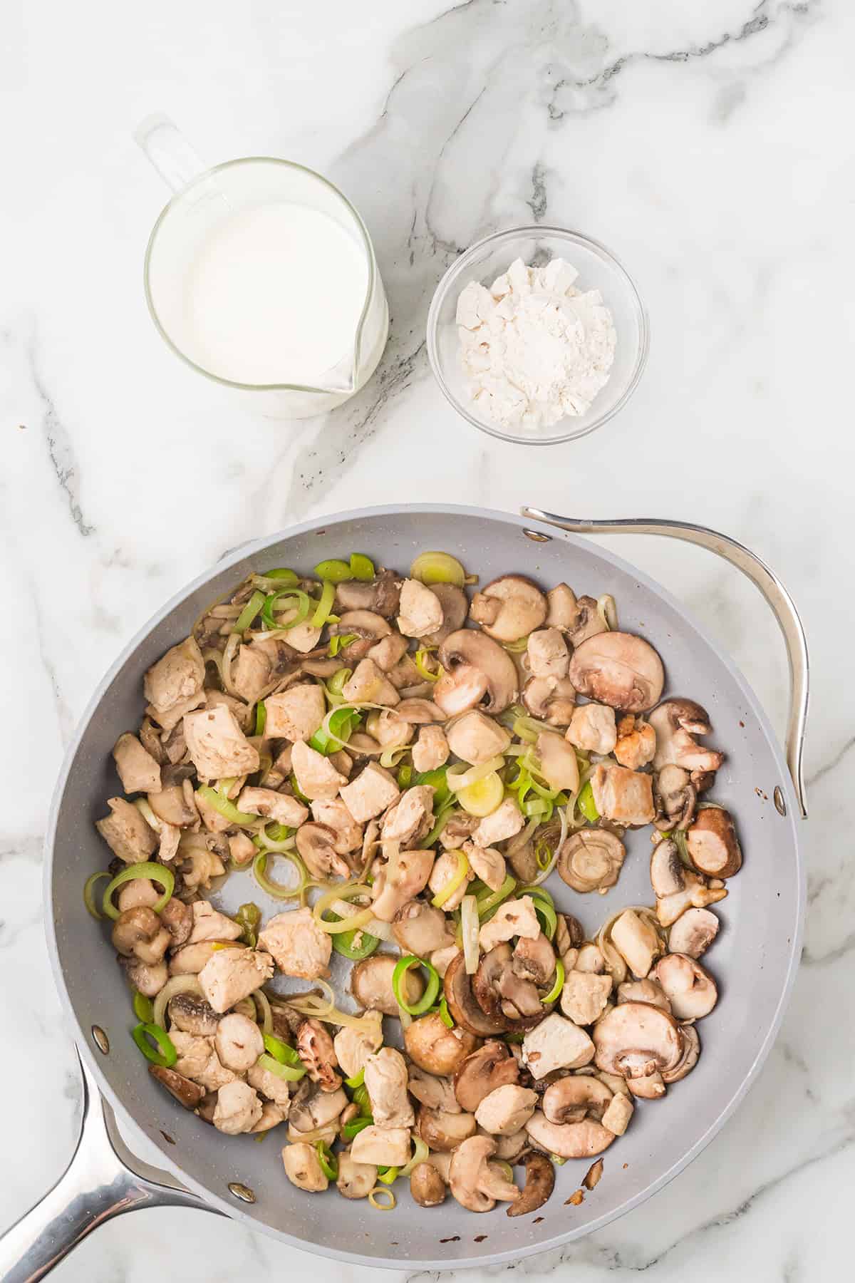 A pan with cooked chicken, mushrooms, and leeks, next to a cup of milk and a bowl of flour on marble.