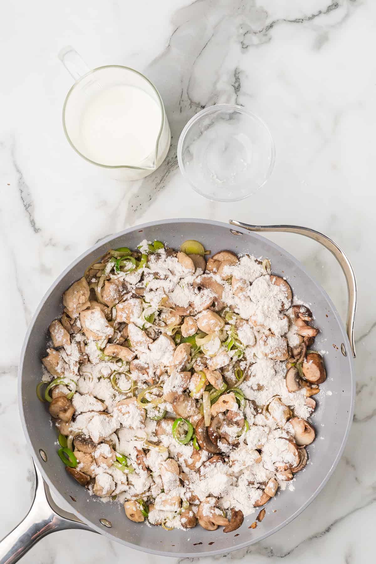 A skillet with mushrooms, leeks, and flour, next to a cup of milk and a small bowl of water on a marble surface.