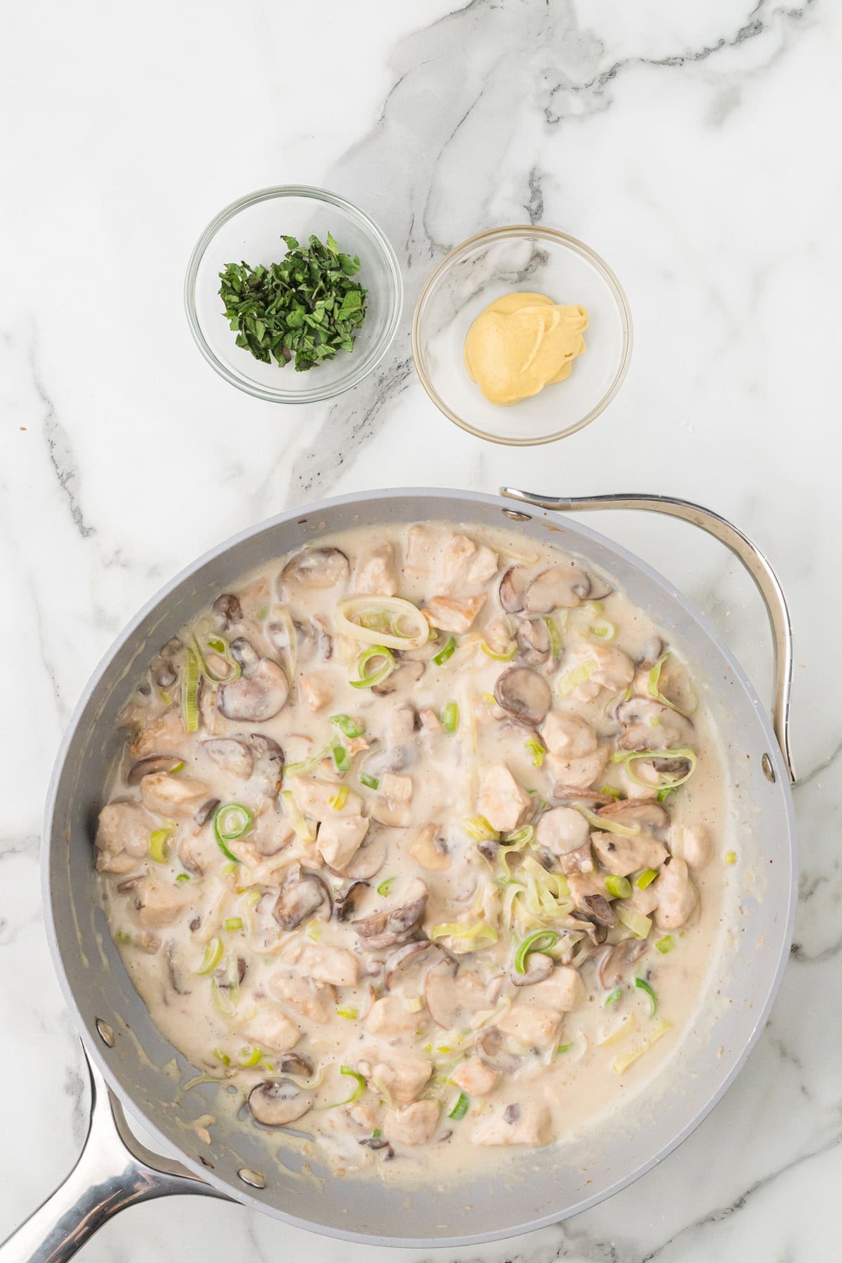 A pan of creamy mushroom and chicken sauce, with bowls of chopped herbs and mustard on the side.