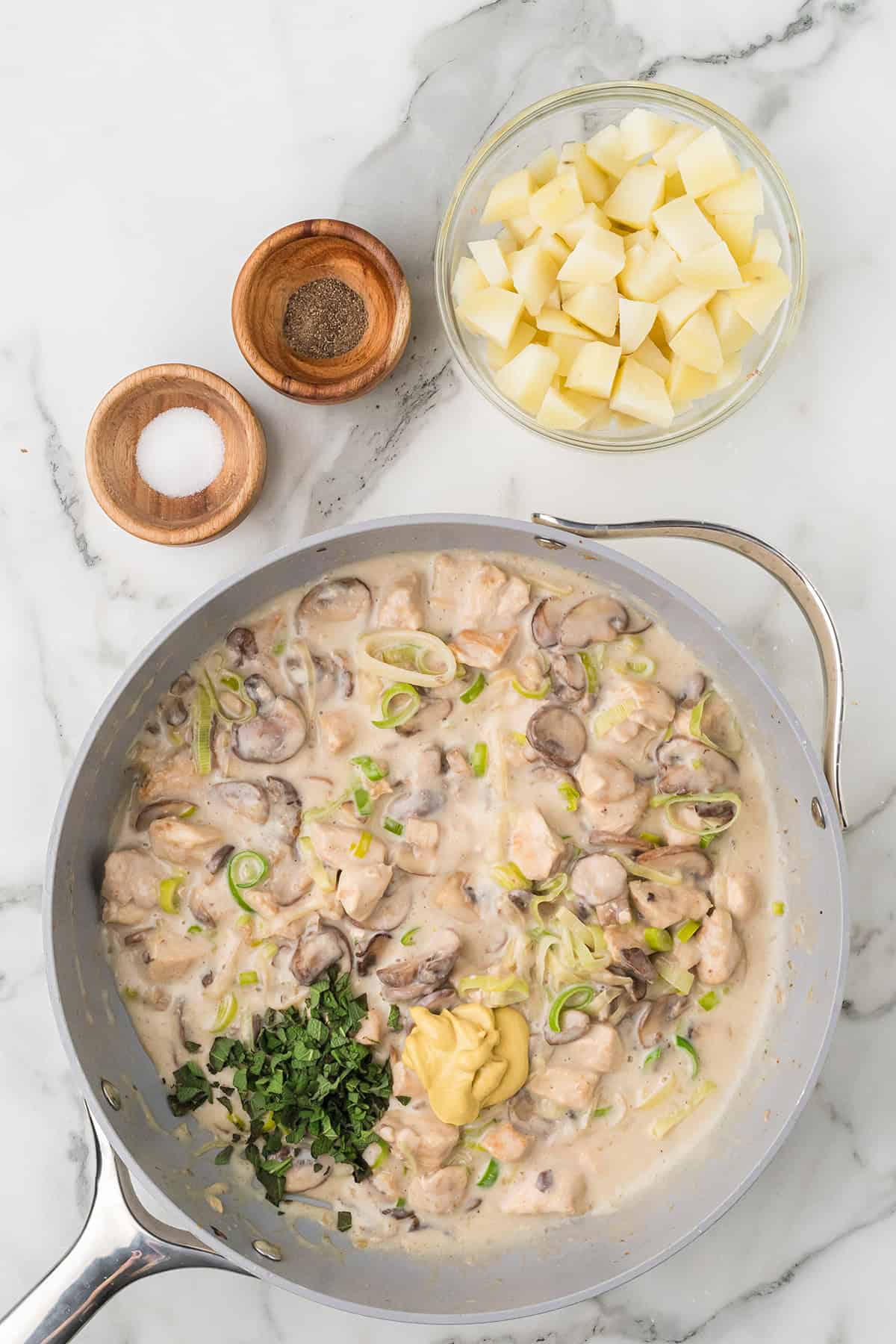 A skillet with creamy mushroom chicken, fresh herbs, mustard, and a bowl of cubed potatoes on a marble countertop.