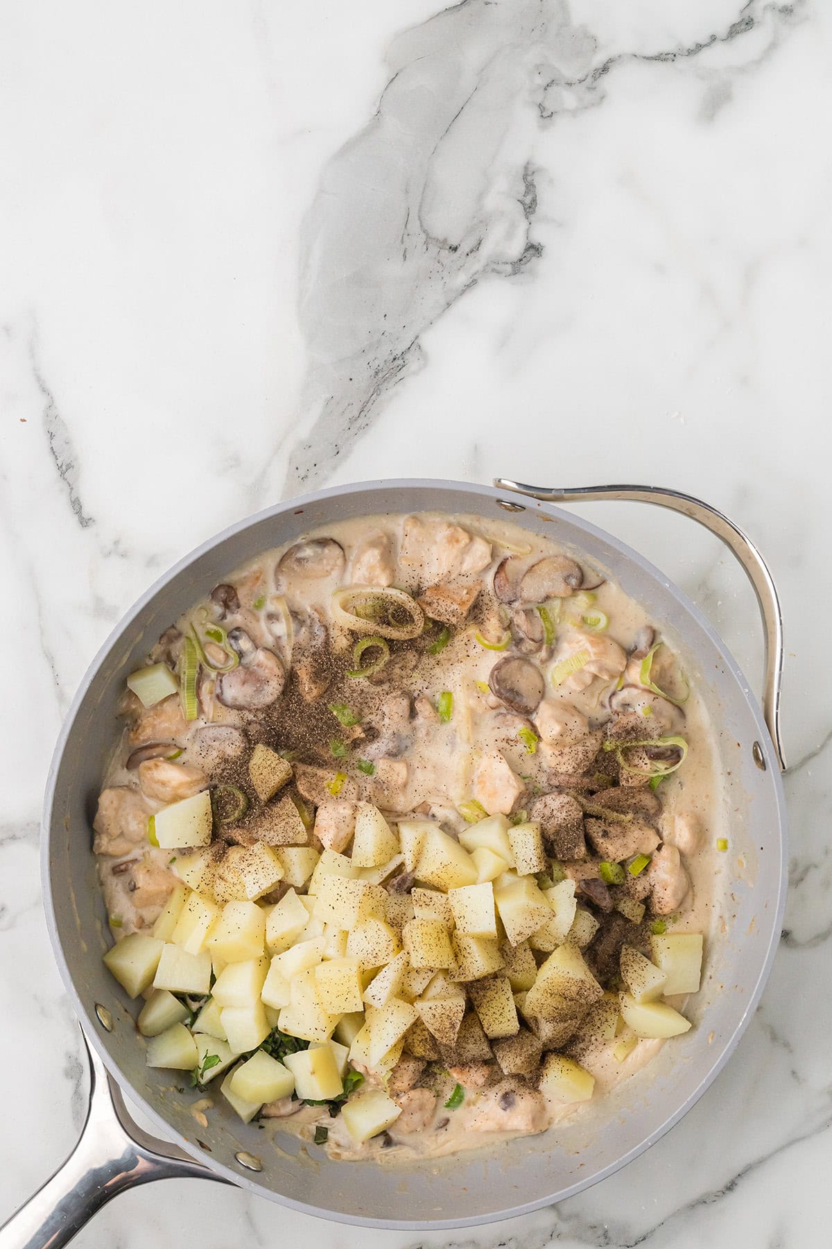 A skillet with cubed potatoes, mushrooms, meat, and creamy sauce on a marble countertop.