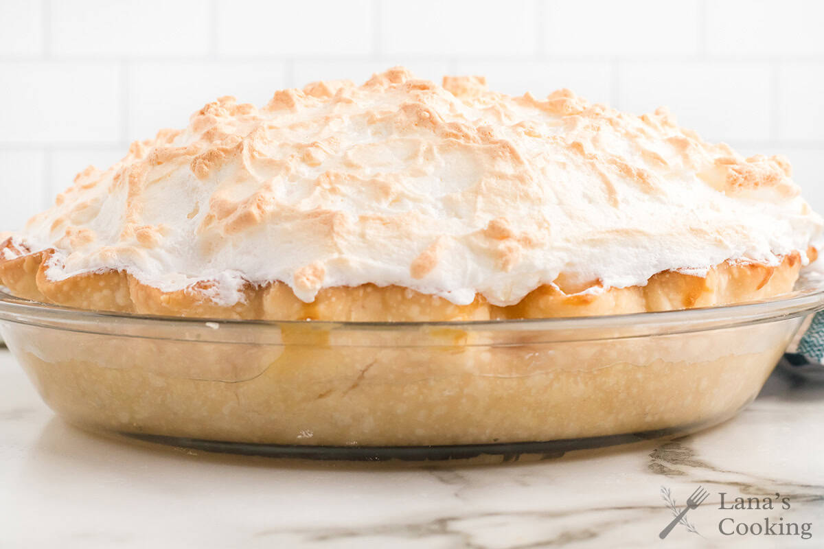 A lemon meringue pie with golden, fluffy meringue topping in a glass pie dish on a marble countertop.