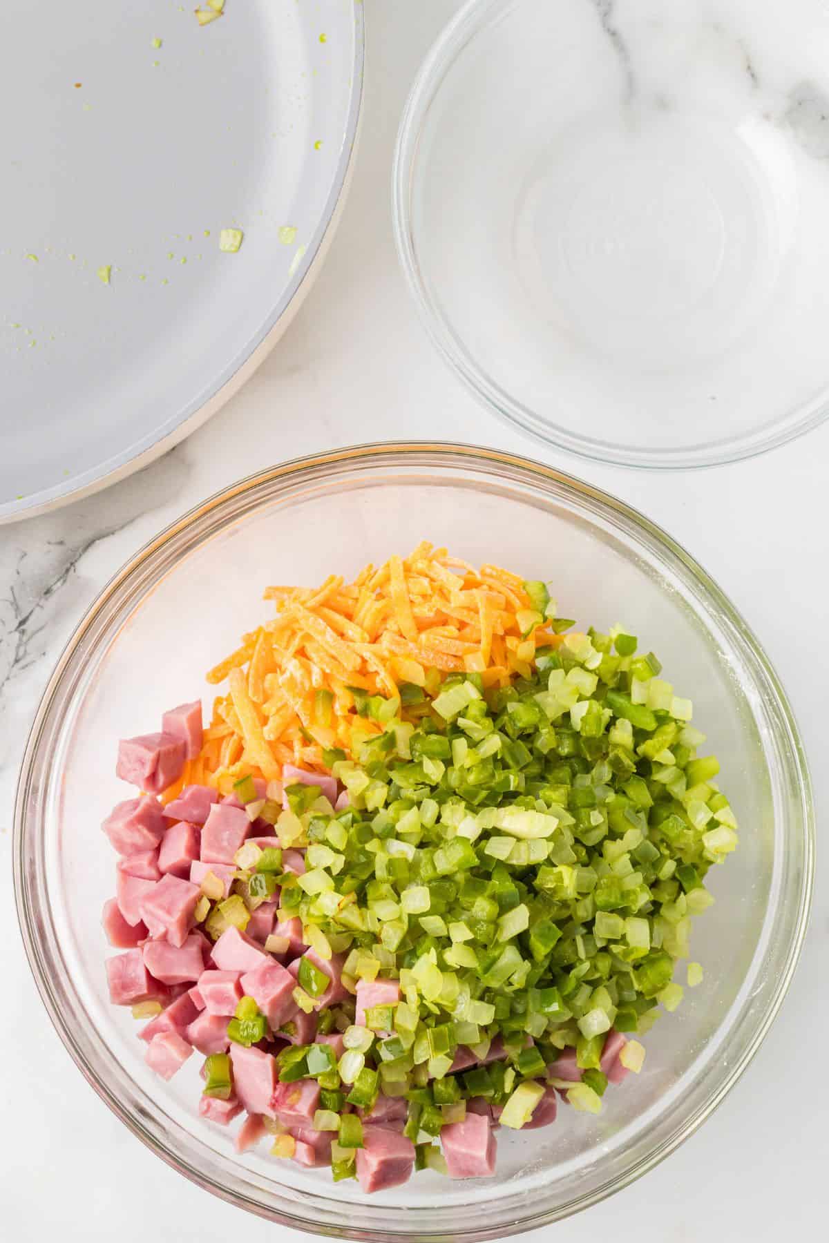 Cheese, diced ham, and the green pepper onion mixture in a glass bowl before being mixed.