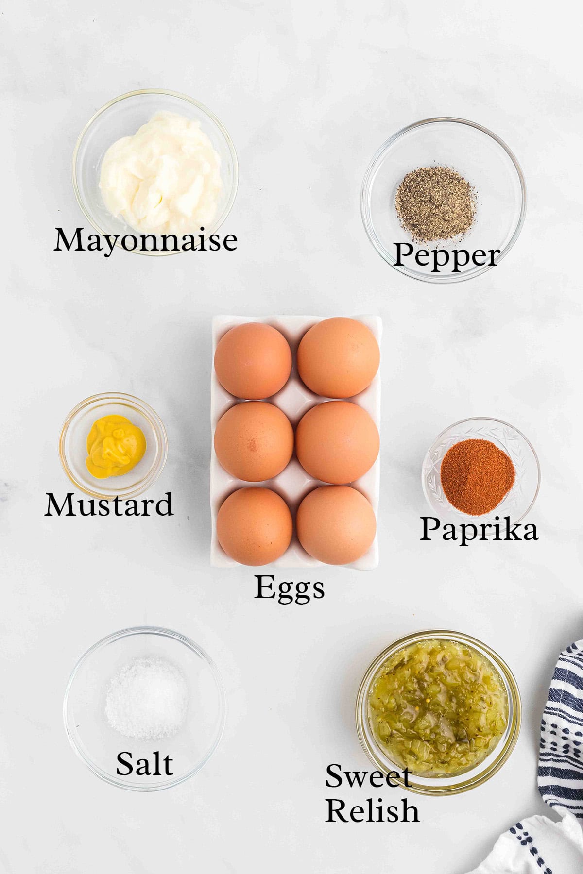 Seven labeled bowls with eggs, mayonnaise, mustard, salt, pepper, paprika, and sweet relish on a white surface.