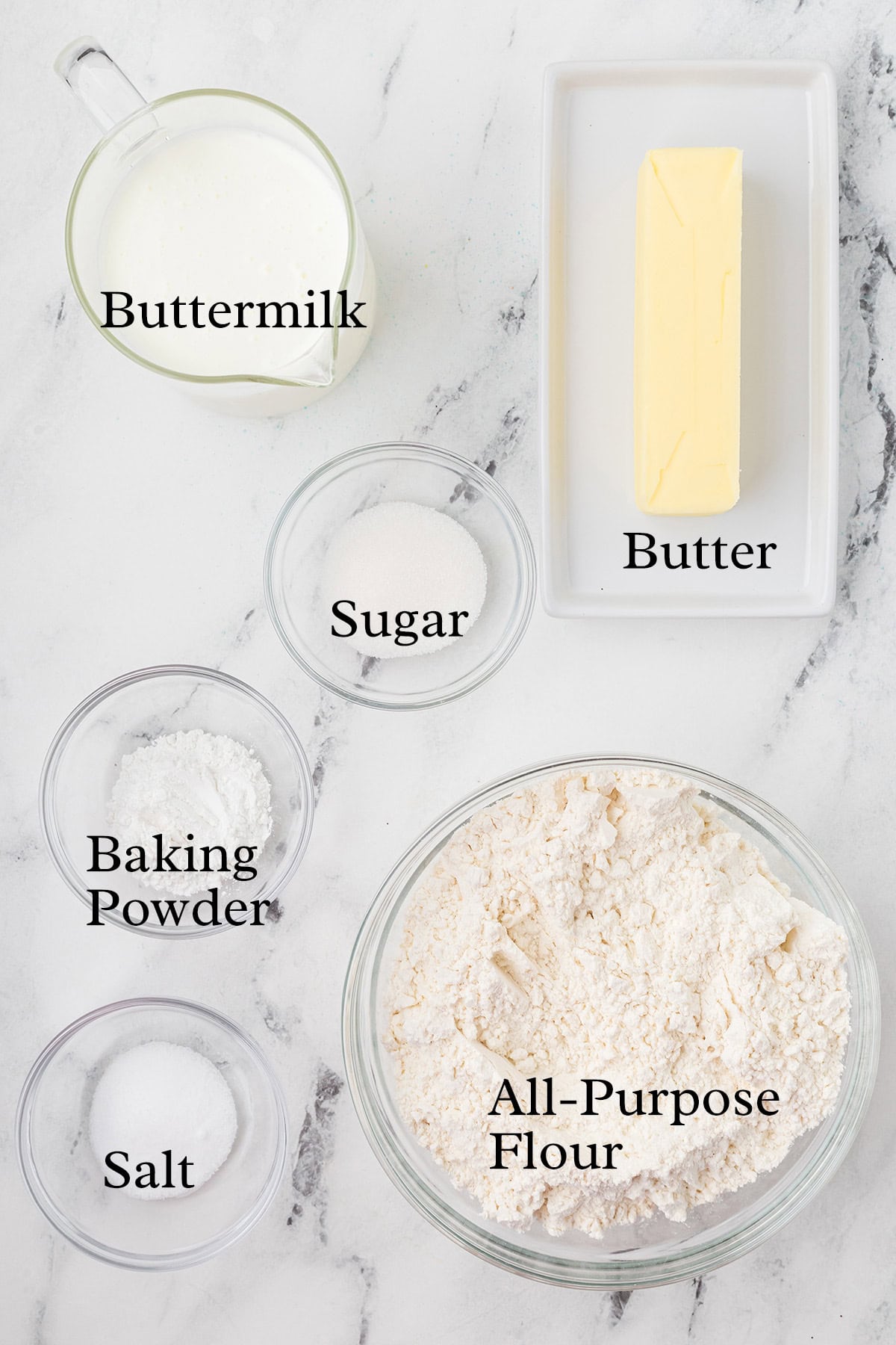 Baking ingredients on a counter: buttermilk, butter, sugar, baking powder, salt, and all-purpose flour in bowls.