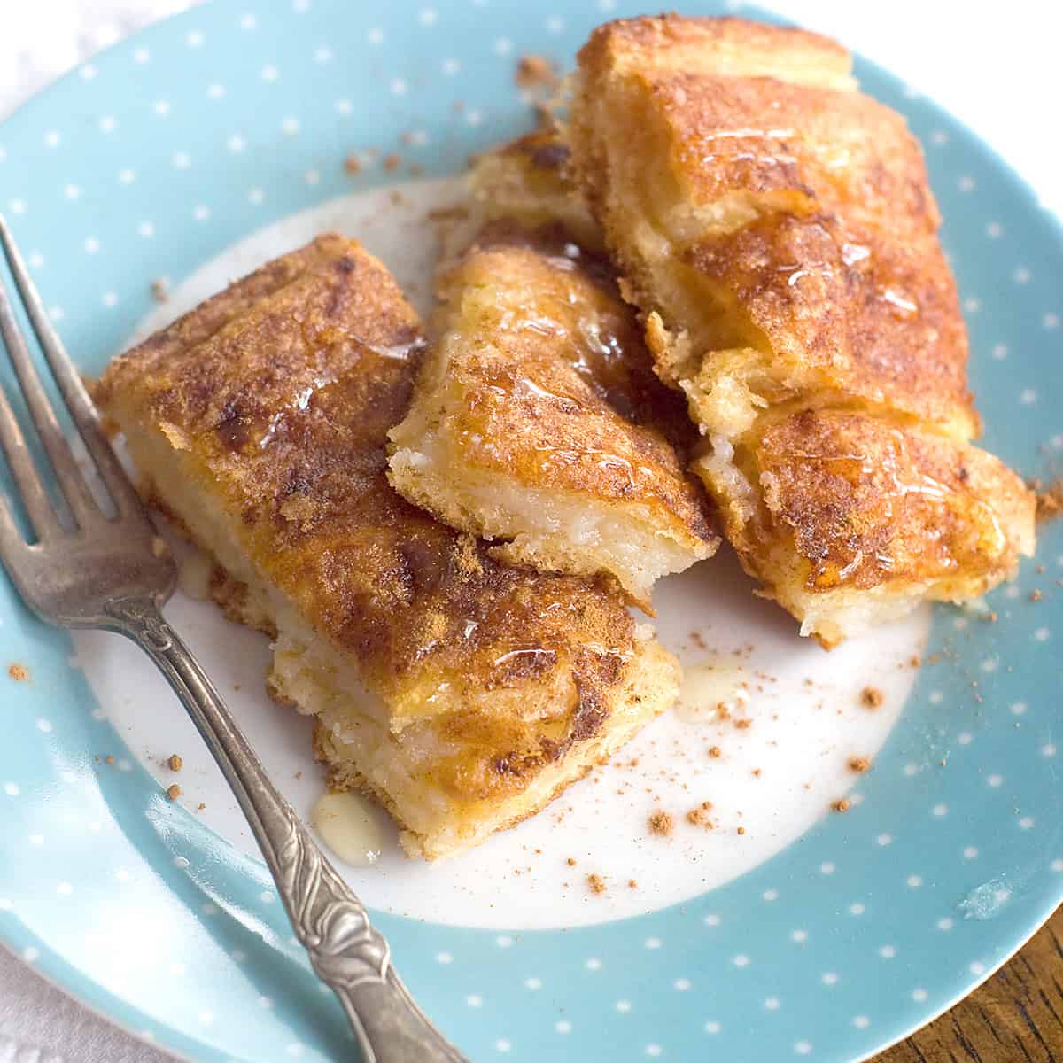 Three pieces of golden-brown cream cheese filled crescent roll pastries with visible cinnamon, served on a light blue plate with white polka dots, next to a fork.
