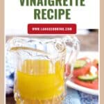 A glass pitcher of lemon vinaigrette next to a plate of salad with tomatoes and feta cheese.