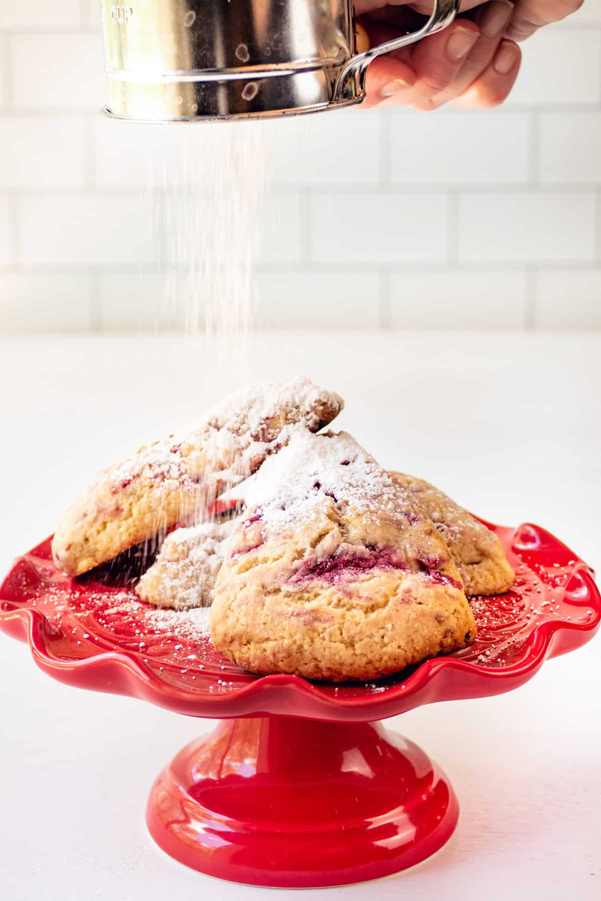 A hand dusts powdered sugar over scones on a red cake stand against a white tiled background.