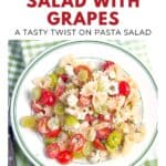 A plate of bow tie pasta salad with grapes, tomatoes, and cheese on a green-checked tablecloth.
