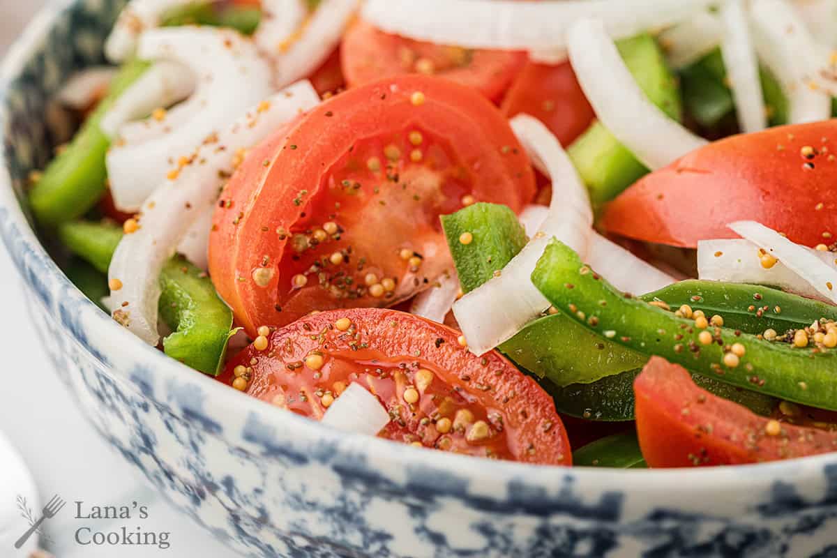 Sliced tomatoes, onions, and green peppers with seasoning in a blue and white bowl.
